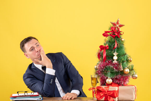 放前视图被贬低的人把他的手放在他的脸颊上坐在圣诞树附近的桌子和黄色的礼物礼物桌子黄色