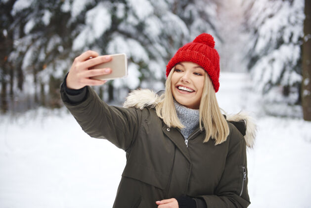 青年微笑的女人在冬天的森林里自拍手机享受文化