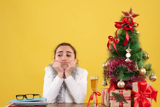 快乐正面图：女医生坐在黄色背景的桌子后面 还有圣诞树和礼品盒坐着人成人