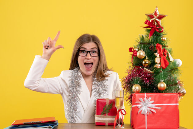 微笑正面图：女医生坐在黄色背景的桌子前 放着圣诞树和礼品盒医生前面女性
