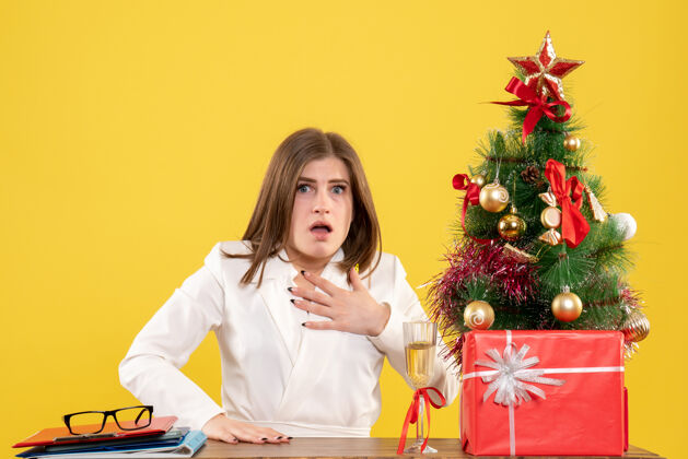办公室正面图：女医生坐在黄色背景的桌子前 放着圣诞树和礼品盒微笑女性快乐