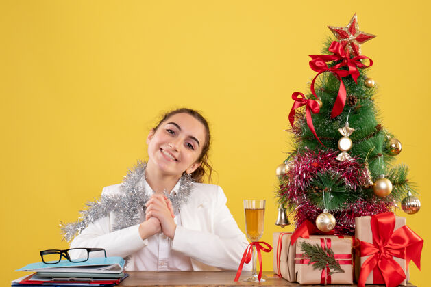 漂亮前视图：女医生坐在黄色背景的桌子后面 带着圣诞树和礼品盒肖像女医生新