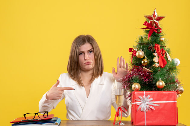 办公室正面图：女医生坐在黄色背景的桌子前 放着圣诞树和礼品盒成人女医生女性