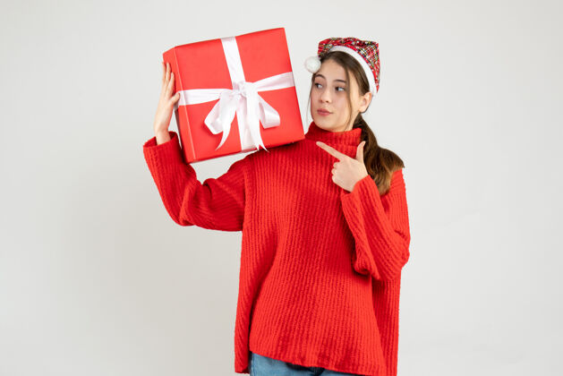 可爱戴着圣诞帽的窥探女孩拿着礼物手指着站在白色地板上的礼物手指礼物圣诞节