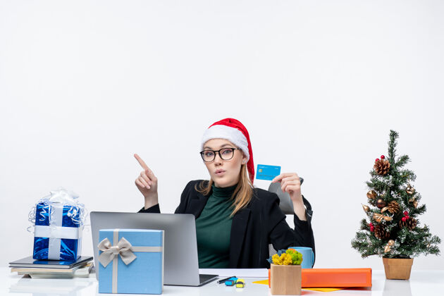 圣诞节一个戴着圣诞老人帽子戴着眼镜的女商人坐在桌子旁 手里拿着圣诞礼物和银行卡坐着戴着商务