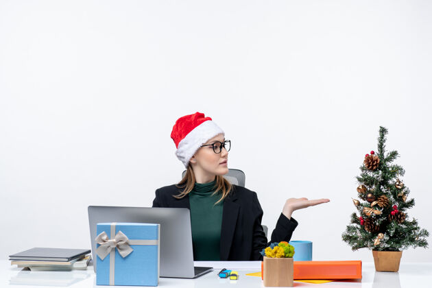 礼物戴着圣诞老人帽的女商人坐在桌子旁 桌上放着圣诞树和礼物工作区笔记本电脑自由职业者