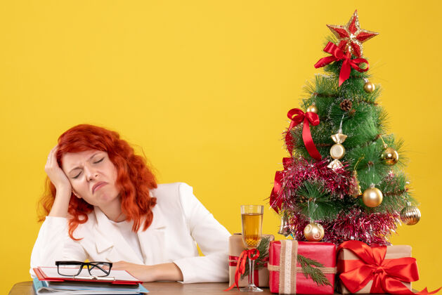 女医生正面图：女医生坐在桌子后面 黄色背景上带着圣诞礼物礼物累了人