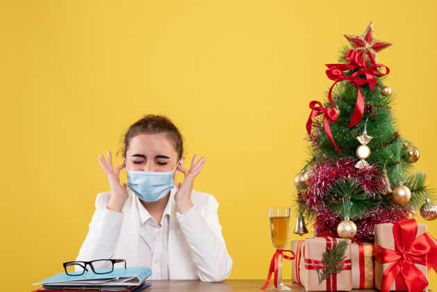 职业正面图女医生戴着防护面具坐在黄色背景上 戴着圣诞树和礼品盒医生男性坐着