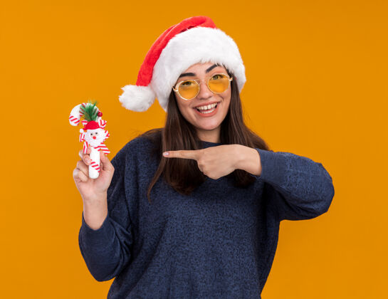 年轻戴着太阳眼镜 戴着圣诞老人帽子的快乐的年轻白人女孩 指着橙色背景上的糖果手杖 还有复制空间点橙色手杖