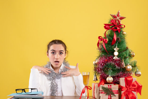 情感前视图：女医生坐在桌子后面 拿着圣诞礼物和黄色背景上的树新郎女医生医生