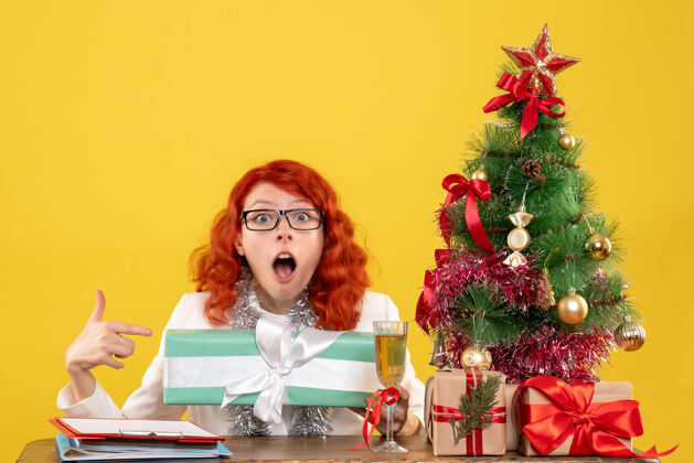 圣诞节前视图：女医生拿着圣诞礼物和黄色背景上的圣诞树坐着医生情感坐着