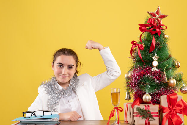 桌子前视图：女医生坐在桌子后面 拿着圣诞礼物和黄色背景上的树弯曲微笑花