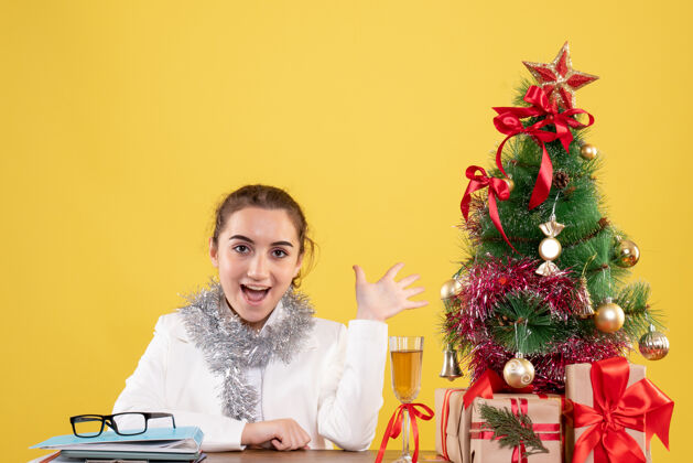 礼物前视图：女医生坐在桌子后面 拿着圣诞礼物和黄色背景上的树树坐着家