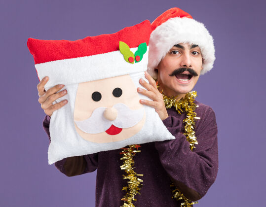 站着一个留着胡子的男人戴着圣诞帽子 脖子上戴着金属丝 手里拿着圣诞枕头 站在紫色的背景上惊讶地看着摄像机惊讶圣诞老人快乐