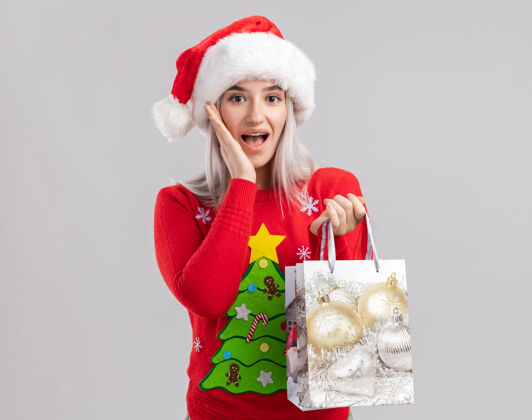 毛衣穿着圣诞毛衣 戴圣诞帽的金发女郎手里拿着装有圣诞礼物的纸袋 看上去既惊讶又惊讶惊讶站着纸