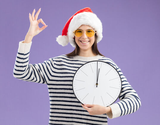 手势戴着太阳眼镜 戴着圣诞帽 拿着时钟 打着ok手势的年轻高加索女孩微笑着圣诞微笑新年