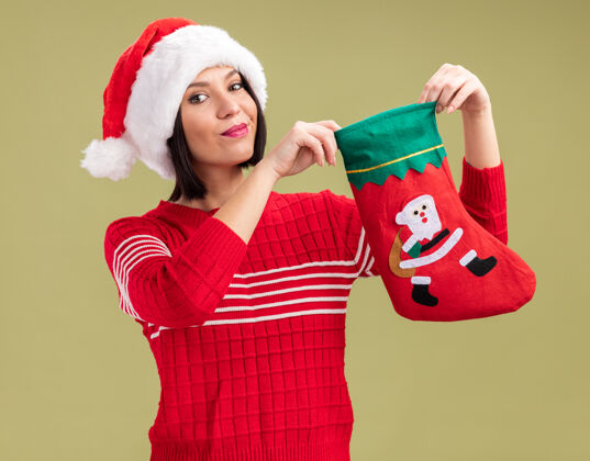 高兴高兴的年轻女孩戴着圣诞帽拿着圣诞长袜孤立在橄榄绿的墙上年轻圣诞老人橄榄