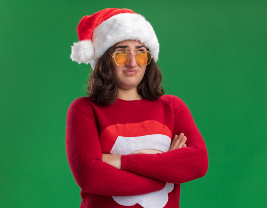 圣诞节穿着圣诞毛衣 戴着圣诞帽和眼镜的年轻女孩困惑地 不高兴地交叉着双臂站在绿色的墙上 望向一边毛衣圣诞老人不高兴