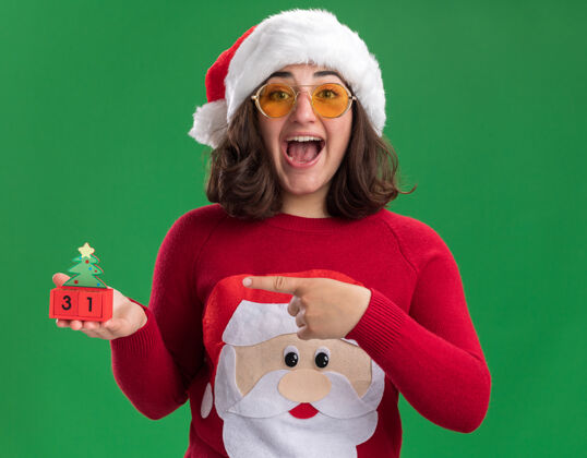 指向穿着圣诞毛衣的小女孩戴着圣诞帽 戴着眼镜 手里拿着新年礼物的玩具立方体 用食指指着它微笑着 站在绿色的墙上 快乐而兴奋站着索引圣诞老人