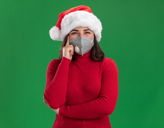 圣诞老人穿着红毛衣 戴着圣诞帽 戴着护面面具的年轻女孩站在绿色的墙上沉思着面部穿着圣诞节