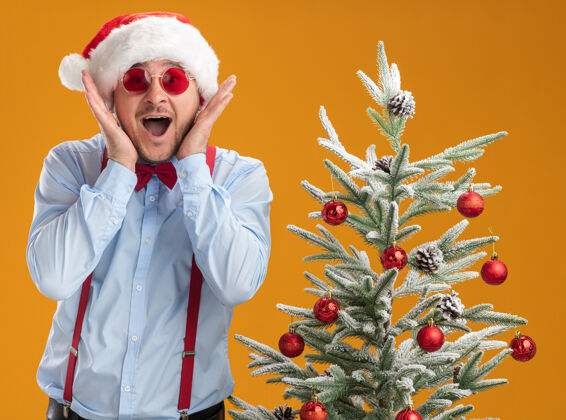 惊讶站在圣诞树旁的那个戴着圣诞帽 戴着红眼镜 系着吊带 打着领结的年轻人 在橘色的墙上惊讶不已惊讶吊带圣诞老人