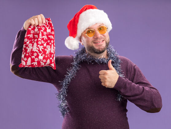 圣诞老人微笑的中年男子戴着圣诞帽 脖子上戴着金箔花环 戴着眼镜 手里拿着圣诞礼品袋 在紫色的墙上孤立地竖起大拇指穿着周围花环