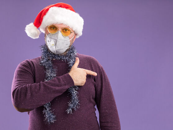侧面令人印象深刻的中年男子戴着圣诞帽 戴着防护面具 脖子上戴着金属丝花环 眼镜指向一侧 隔离在紫色墙壁上 留有复制空间印象深刻周围穿着