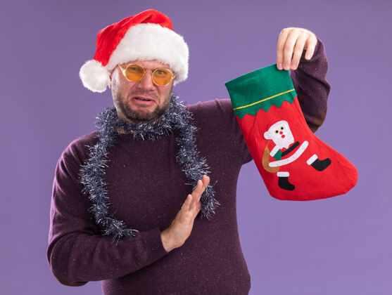 脖子一个戴着圣诞帽 脖子上戴着金箔花环 戴着眼镜 手里拿着圣诞长袜的中年男人 在紫色的墙上做着拒绝的手势长袜穿拒绝