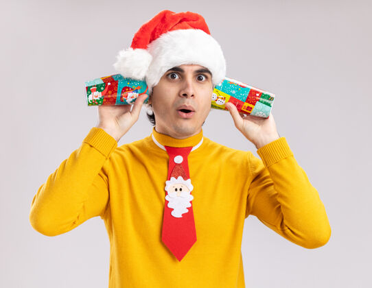 领带一个穿着黄色高领毛衣 戴着圣诞帽 打着滑稽领带的年轻人站在白色的墙上 手里拿着纸杯捂着耳朵 看上去很困惑圣诞节站着帽子