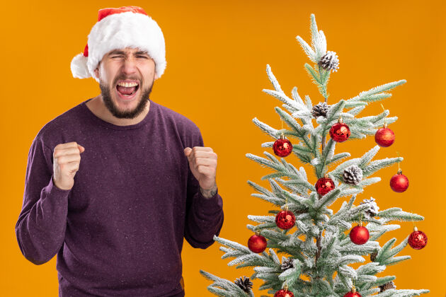 毛衣身穿紫色毛衣 头戴圣诞帽的年轻人握紧拳头 站在橙色背景下的圣诞树旁 气势汹汹地大喊大叫男人喊叫圣诞树