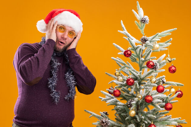 花环惊讶的中年男子戴着圣诞帽 脖子上戴着金箔花环 戴着眼镜 站在装饰好的圣诞树旁 手放在头上 孤立地站在橙色的墙上保持穿着圣诞节