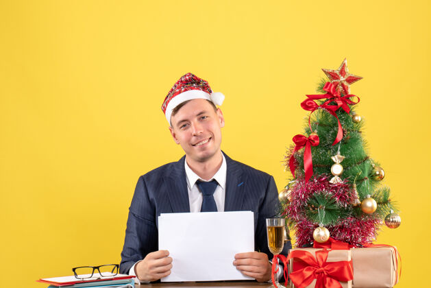 办公室圣诞树旁坐着戴着圣诞帽的快乐男人的正面图 黄色的礼物桌子黄色坐着