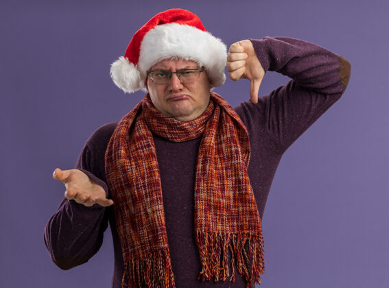 眼镜皱眉的成年男子戴着眼镜 戴着圣诞帽 脖子上围着围巾 看着摄像机 镜头里空手而下 拇指被隔离在紫色背景上皱眉成人空
