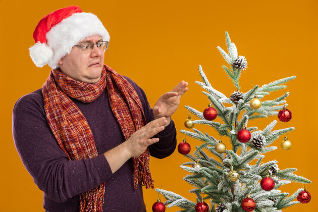 围巾戴着眼镜 戴着圣诞帽 脖子上围着围巾 站在装饰好的圣诞树旁 在橙色的背景下看圣诞树快乐做圣诞快乐