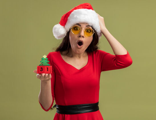 漂亮戴着圣诞帽 戴着眼镜 手持圣诞树玩具 手放在头上 看着橄榄绿背景上的相机圣诞树眼镜圣诞快乐