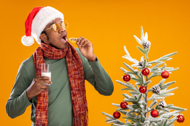 饼干一个戴着圣诞帽 围着围巾 拿着饼干 拿着牛奶的非洲裔美国人站在橙色背景下的圣诞树旁美国人非洲围巾