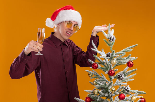 快乐戴着圣诞帽 戴着圣诞眼镜的快乐的金发年轻人站在装饰好的圣诞树旁 指着圣诞树 手里拿着一杯香槟 看着孤立在橙色背景上的摄像机圣诞树站着拿着