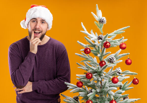 圣诞老人穿着紫色毛衣 戴着圣诞帽的快乐的年轻人看着相机 脸上挂着微笑 旁边是橙色背景下的圣诞树男人看站着