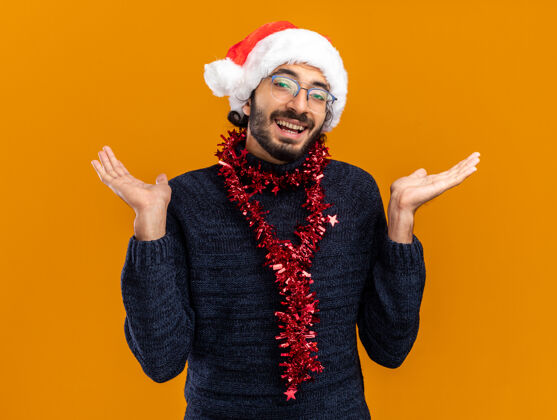 年轻微笑着的年轻帅哥戴着圣诞帽 脖子上戴着花环 两手分开放在橙色的背景上帅气手蔓延