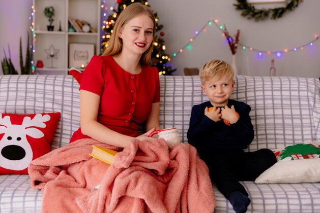 树在一间以圣诞树为背景的装饰房间里 穿着红色连衣裙的快乐妈妈端着一杯茶 她的孩子在毯子下抱着桔子妈妈杯子房间