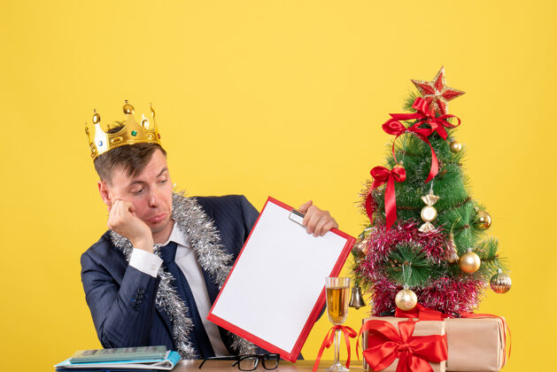 皇冠前视图的商人与皇冠检查文件坐在桌子旁圣诞树和黄色的礼物商务圣诞节树