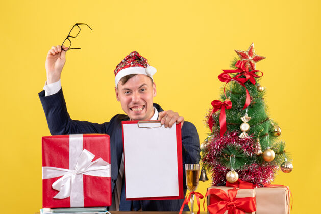 坐着正面图：快乐的商人摘下眼镜坐在圣诞树旁的桌子旁 黄色的礼物眼镜男人礼物