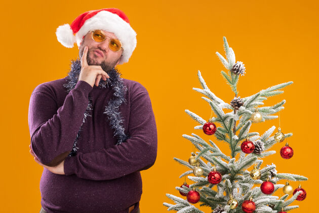 树体贴的中年男子戴着圣诞帽 脖子上戴着金箔花环 戴着眼镜 站在装饰好的圣诞树旁圣诞老人中年周围