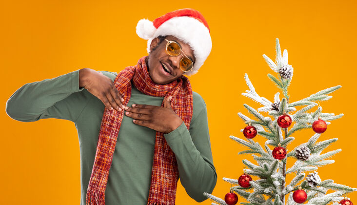 站着戴着圣诞帽 围着围巾 双手放在胸前的非洲裔美国人站在橙色墙上的圣诞树旁 心情愉快 充满感激之情脖子帽子圣诞老人