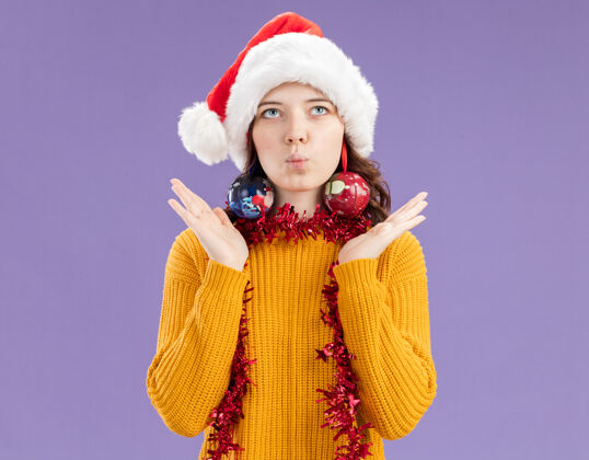 帽子戴着圣诞帽 脖子上戴着花环的年轻斯拉夫女孩高兴地把玻璃球饰物戴在耳朵上 背景是紫色 有复制空间玻璃花环耳朵