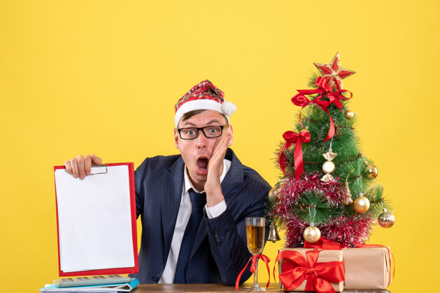 西装前视图震惊的人拿着剪贴板坐在圣诞树附近的桌子上 黄色的礼物职业树圣诞节