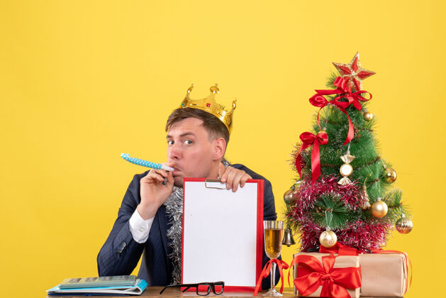 坐着前视图的商人与皇冠使用noisemaker坐在桌旁圣诞树和黄色的礼物男性管乐器生意