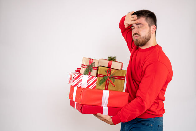 快乐身穿红衫的年轻人拿着圣诞礼物 在白墙上感到疲倦男前面年轻