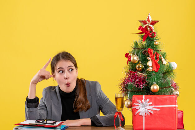 展示前视图惊讶的女孩坐在桌旁展示圣诞树和鸡尾酒礼物一些东西女孩树