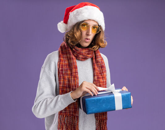 站着戴着圣诞老人帽 戴着黄色眼镜 脖子上围着暖和的围巾的年轻人手里拿着圣诞礼物 站在紫色背景下焦急地看着相机围巾眼镜帽子